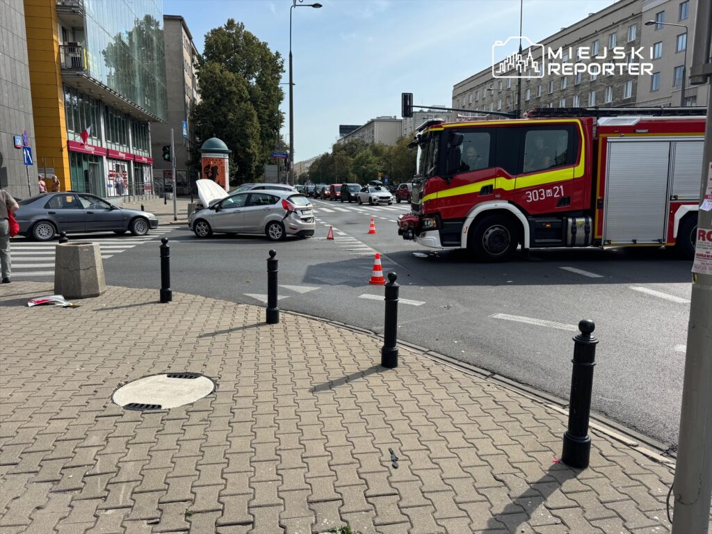 W miejscu zdarzenia drogowego stoi wóz strażacki, a na jezdni widoczne są uszkodzone samochody i pachołki ostrzegawcze.