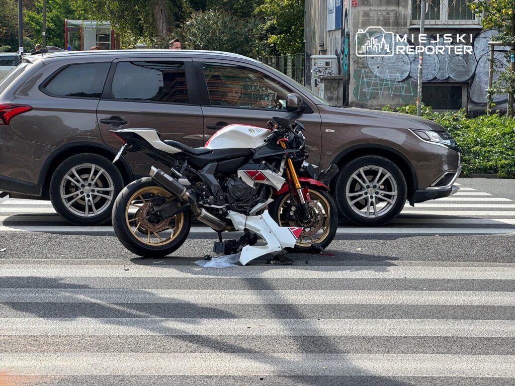 Motocykl leży na pasach, obok zatrzymanego samochodu osobowego, w miejskim otoczeniu z graffiti w tle.
