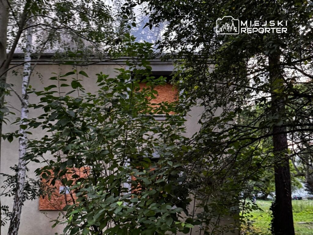 Zarośnięty budynek z zasłoniętymi oknami, otoczony gęstą roślinnością w miejskim otoczeniu.