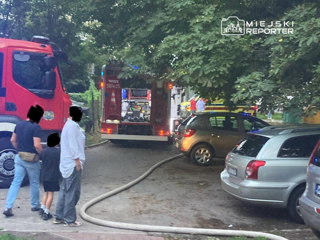 Grupa osób obserwuje działania straży pożarnej przy zaparkowanych samochodach na wąskiej ulicy.
