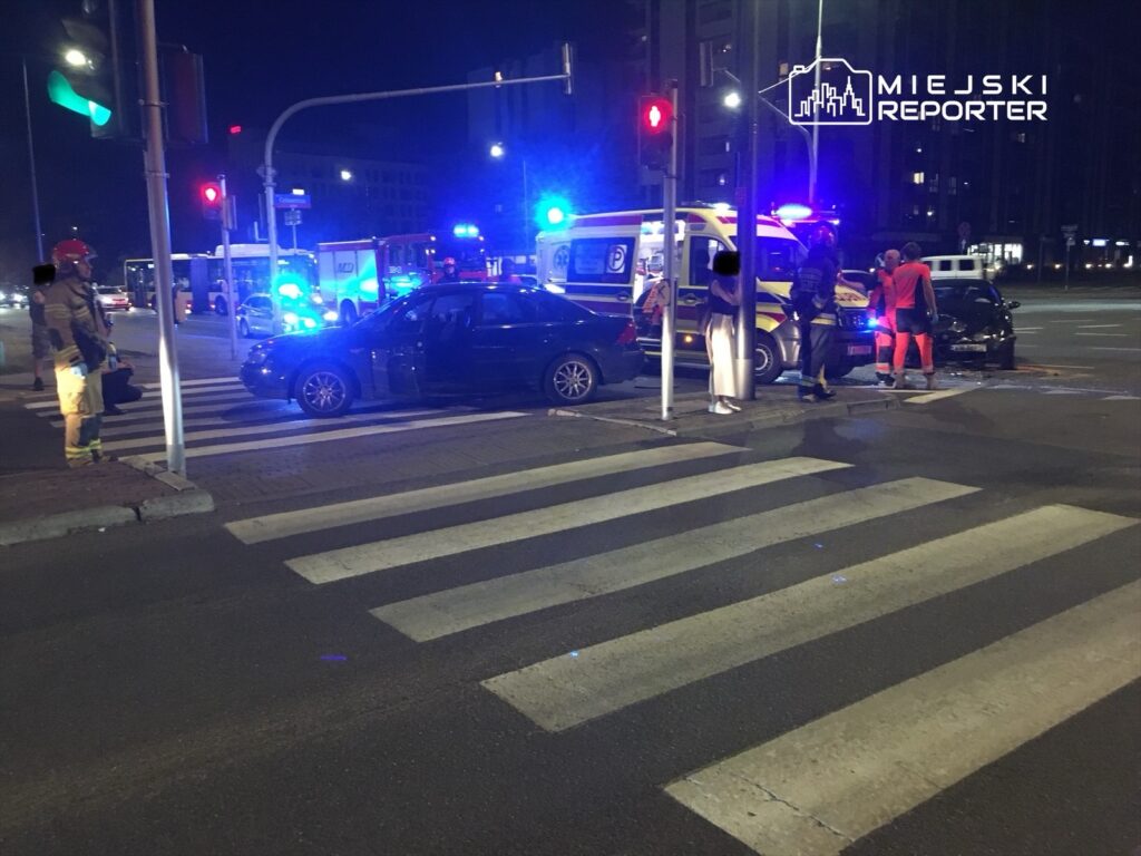 Na skrzyżowaniu w nocy, ratownicy medyczni i strażacy udzielają pomocy poszkodowanym po wypadku drogowym.