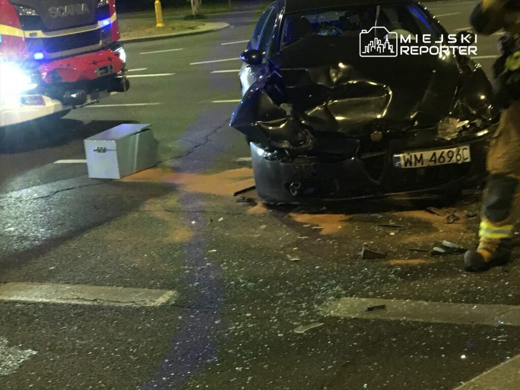 Zniszczony czarny samochód osobowy z wgniecionym przodem stoi na drodze, obok widoczna skrzynka i resztki szkła.