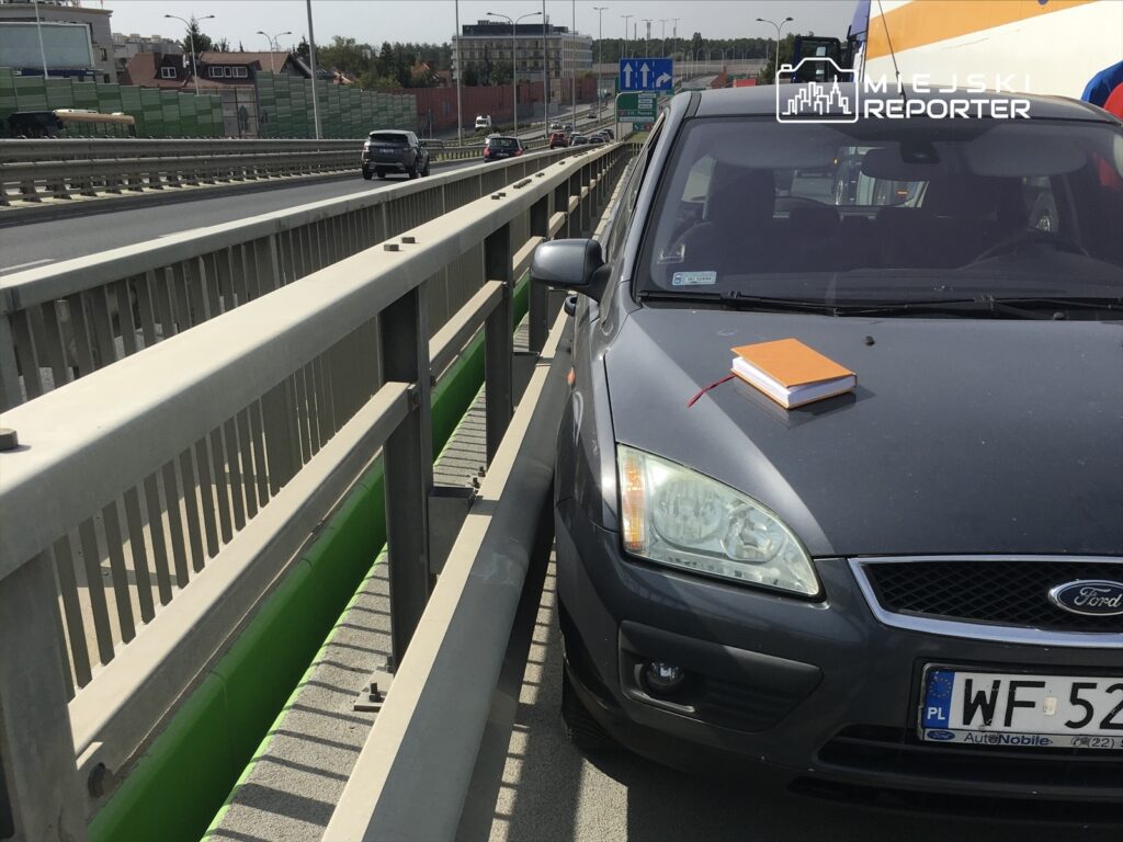 Samochód osobowy stojący na pasie ruchu z pomarańczowym notatnikiem na masce, otoczony barierkami.
