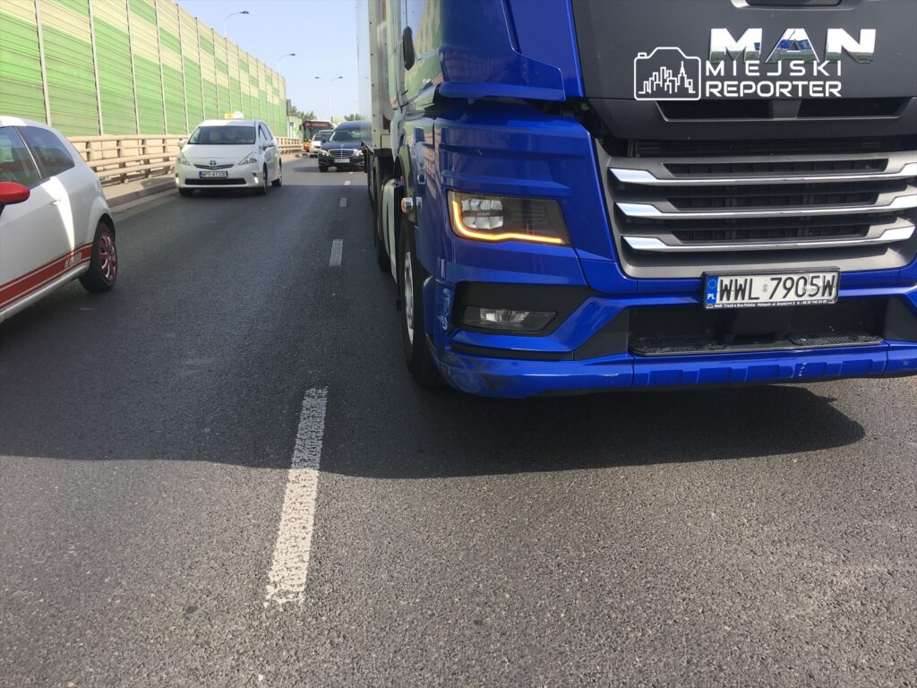 Niebieska ciężarówka MAN zbliża się do innych pojazdów na ruchliwej drodze miejskiej, otoczonej zielonymi ekranami akustycznymi.