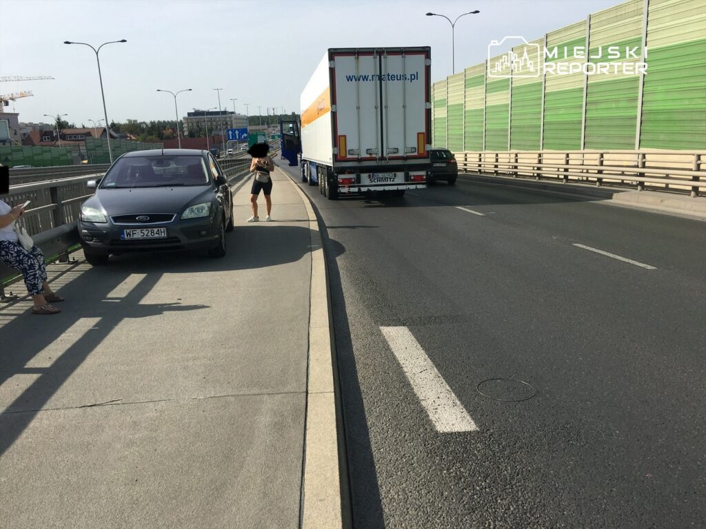 Kobieta stoi na chodniku obok zaparkowanego samochodu, podczas gdy ciężarówka przejeżdża obok na drodze.