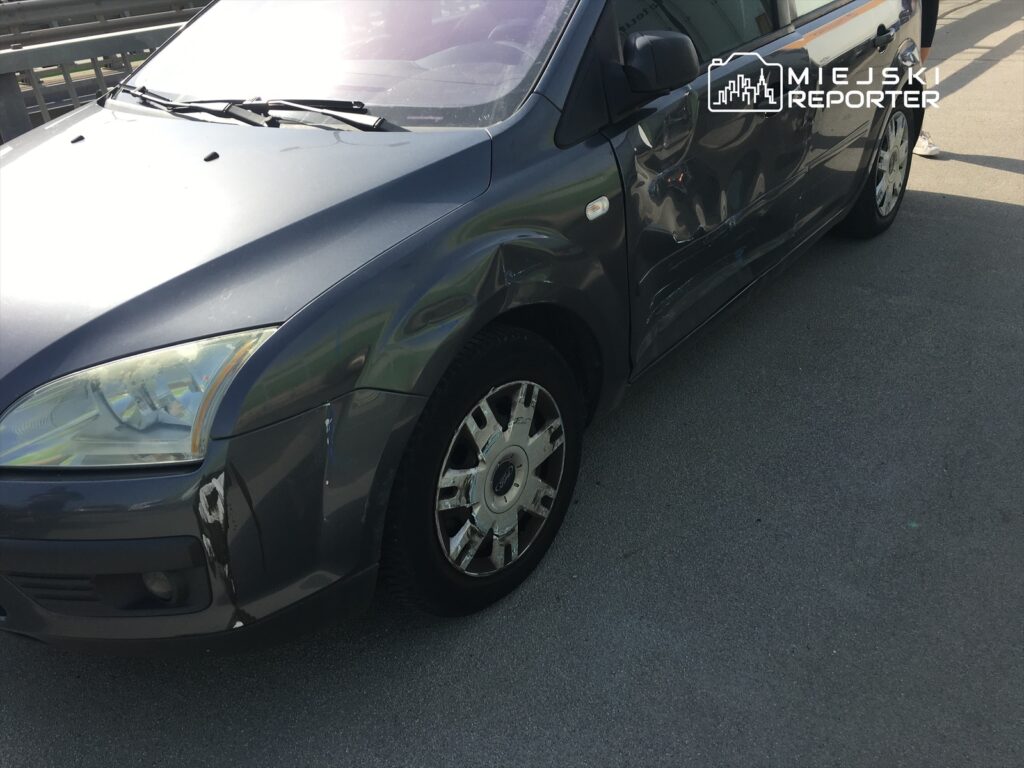 Uszkodzony szary samochód osobowy zaparkowany na asfaltowej nawierzchni, z widocznymi zarysowaniami na karoserii.