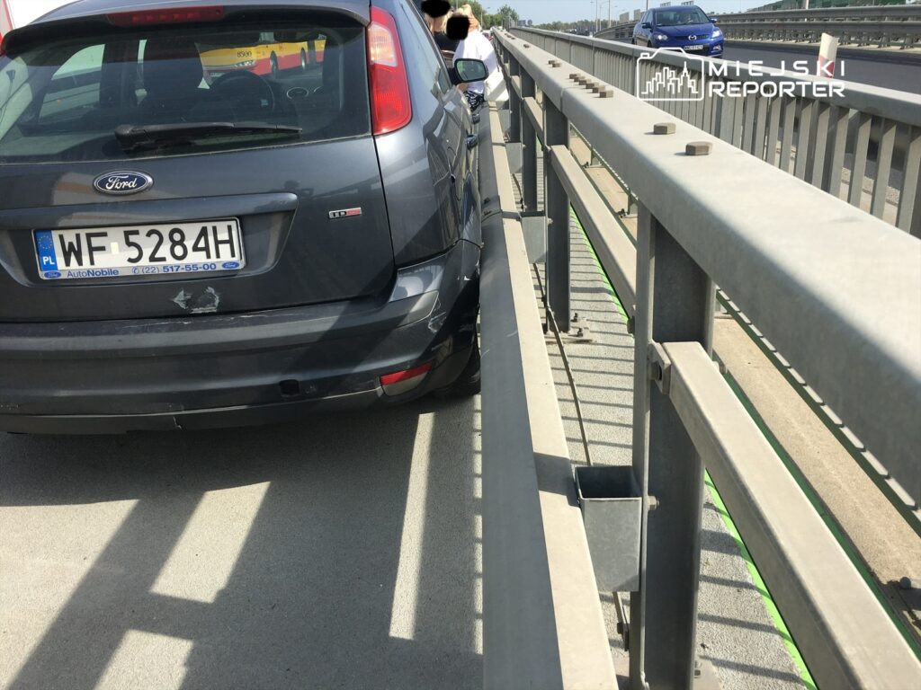 Czarny samochód osobowy zaparkowany blisko metalowej bariery na moście, w tle widoczne inne pojazdy.