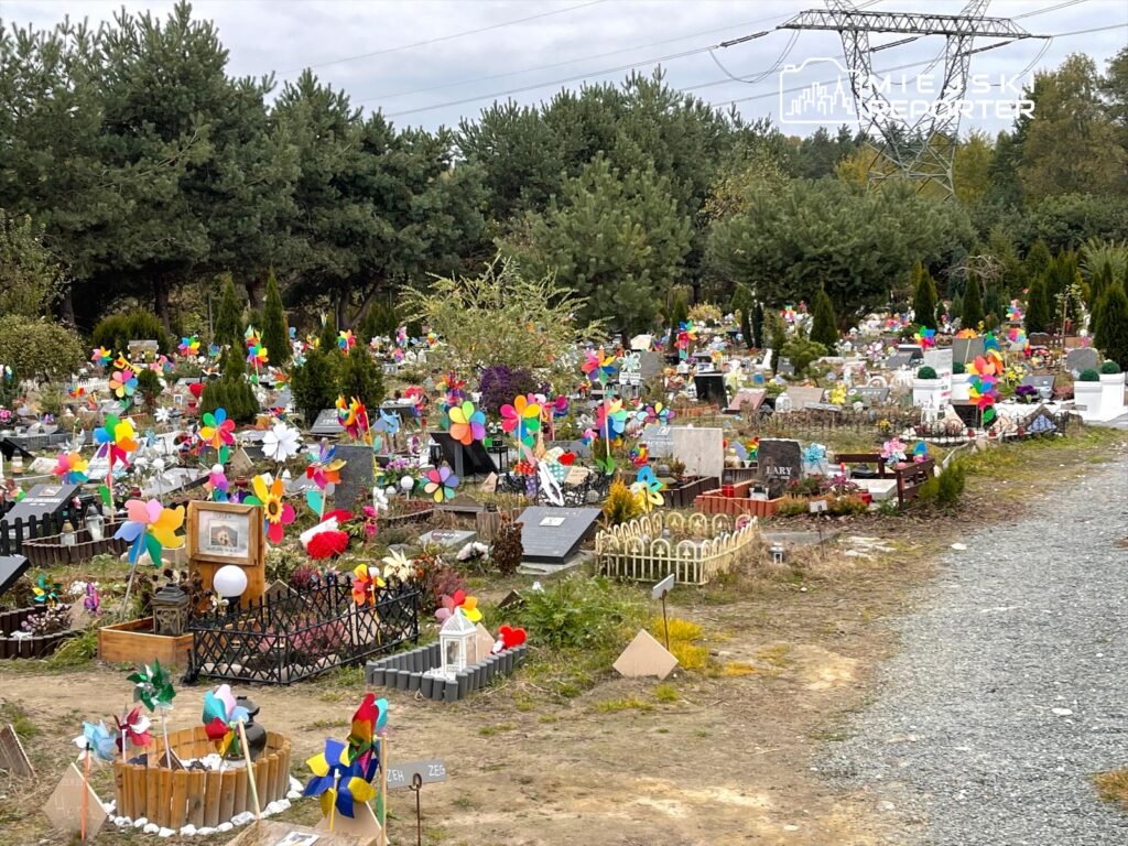 Kolorowe wiatraczki i kwiaty ozdabiają groby na cmentarzu otoczonym drzewami, wśród gęstej zieleni.