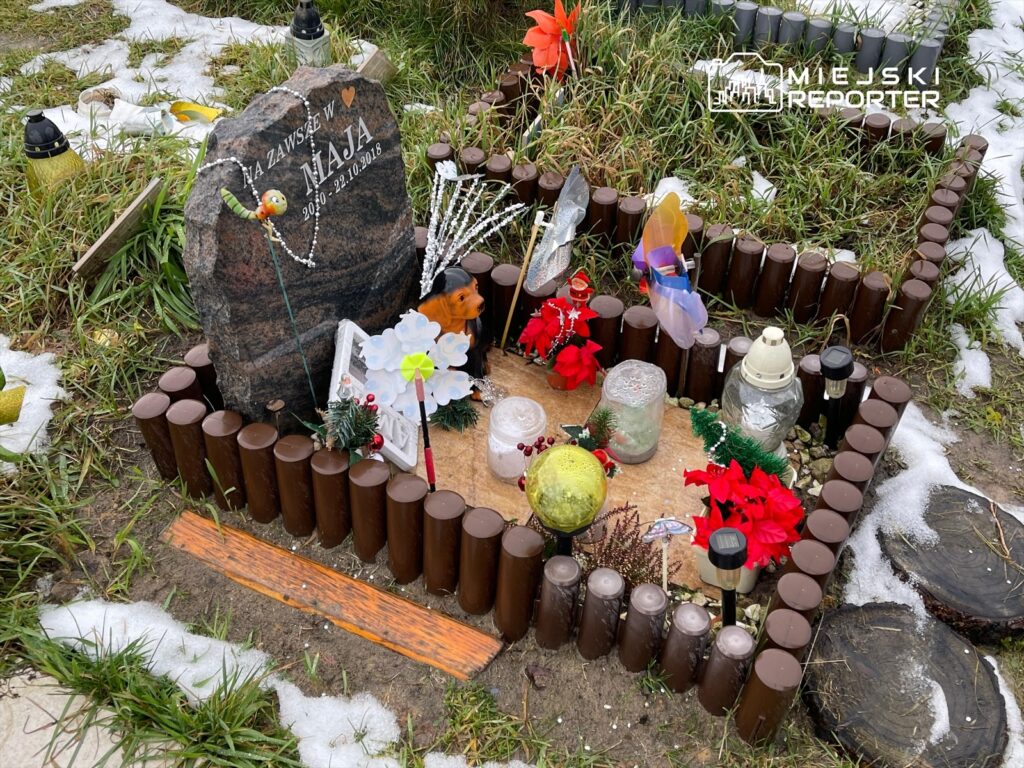 Nagrobek z napisem "NA ZAWSZE W MAJA 2010 - 22.10.2018" otoczony dekoracjami i zniczami na cmentarzu.