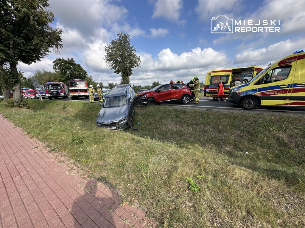 Dwa pojazdy osobowe po wypadku drogowym na poboczu, z interweniującymi strażakami i zespołem ratownictwa medycznego.