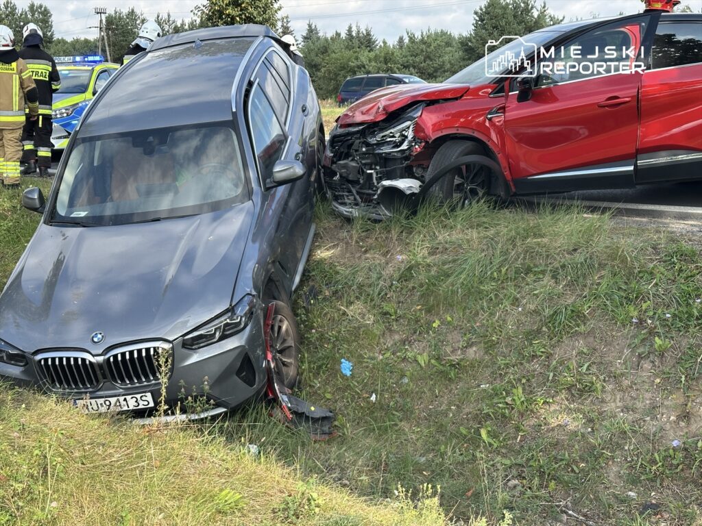 Dwa rozbite samochody osobowe na poboczu drogi, obok stoją strażacy i policjanci.