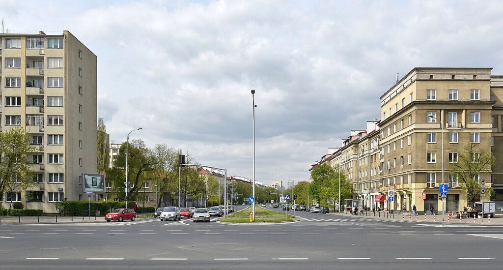 Rondo z ruchem samochodowym, otoczone blokami mieszkalnymi, w tle chmurne niebo.