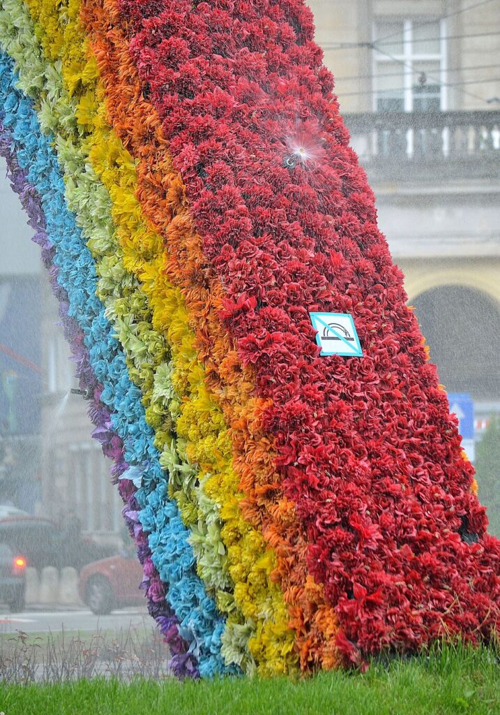 Kolorowa konstrukcja w kształcie tęczy z kwiatów, na tle miasta, spryskiwana wodą.
