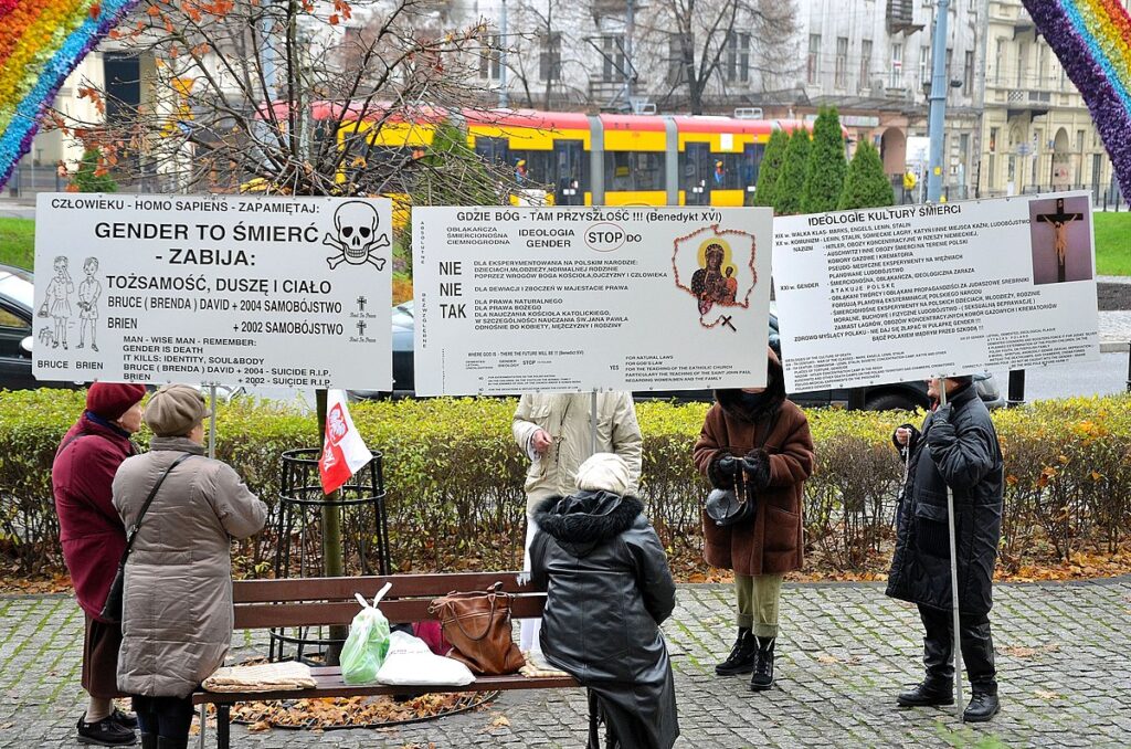 Grupa osób z transparentami protestuje w miejskim parku, w tle widoczny autobus i kolorowa tęcza.