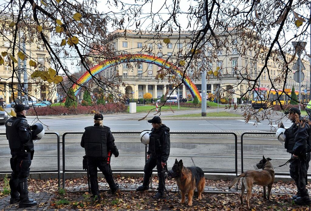 Grupa policjantów w mundurach obserwuje tęczę w miejskim otoczeniu, trzymając psy na smyczach.