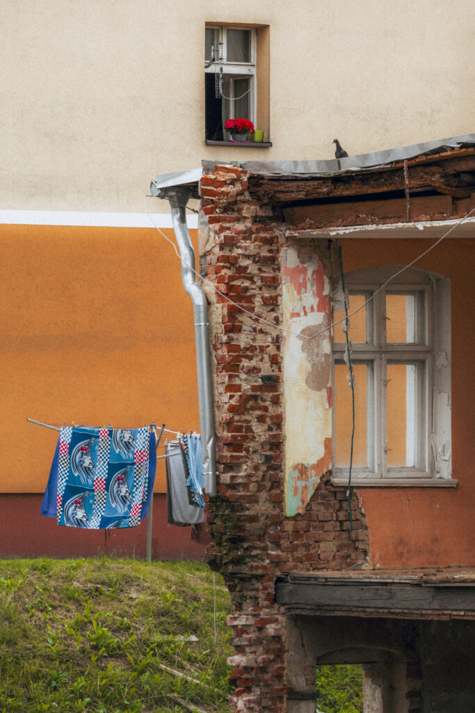 Czerwona ceglana ściana z odsłoniętą cegłą, obok rozwieszone kolorowe pranie na sznurku, w tle okno z kwiatami.