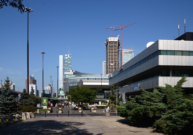 Nowoczesne biurowce w centrum miasta, w tle widoczny plac budowy oraz zieleń w otoczeniu. Jasne niebo bez chmur.
