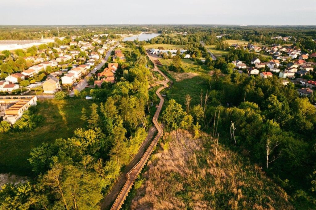 Widok na zespół domów jednorodzinnych otoczonych zielenią, z drewnianą alejką wijącą się przez teren leśny.