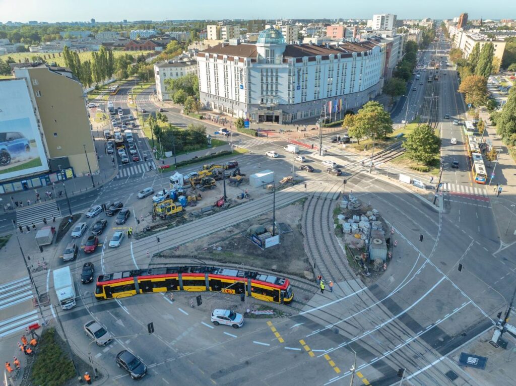 Tramwaj w kolorze czerwono-żółtym skręca na rondzie, otoczony przez ruch samochodowy i prace budowlane.
