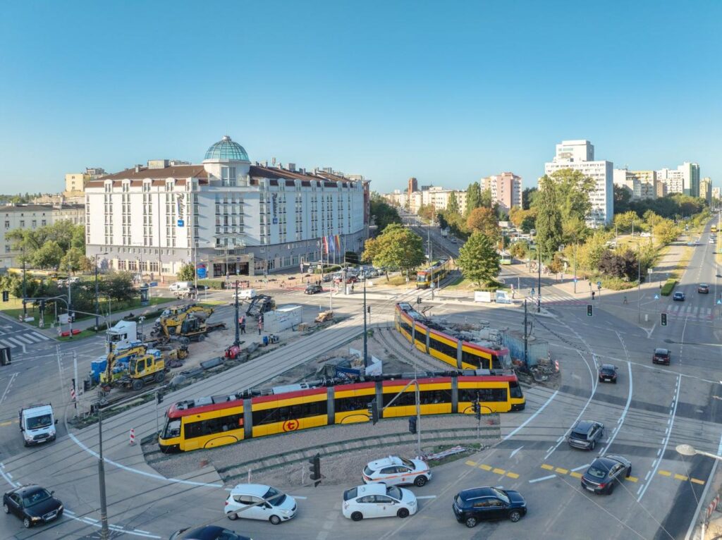 Tramwaj w żółto-czarnych barwach przejeżdża przez skrzyżowanie w miejskiej scenerii z budynkiem hotelowym w tle.