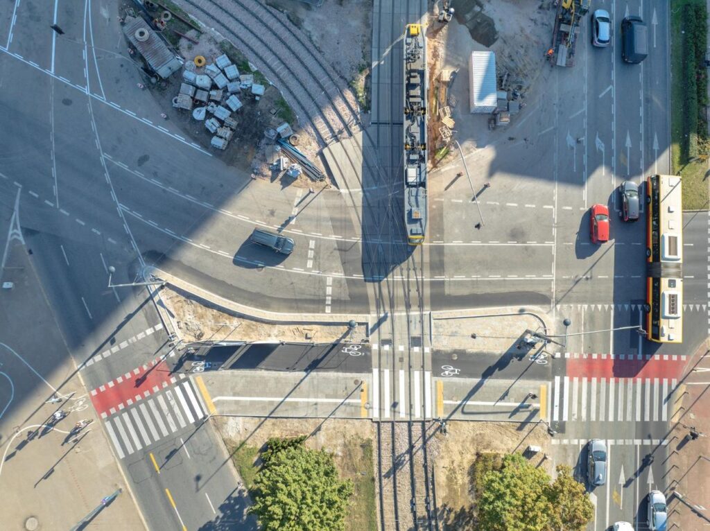 Tramwaj przejeżdża przez skrzyżowanie z nową infrastrukturą rowerową, obok budowy i ruchu ulicznego.