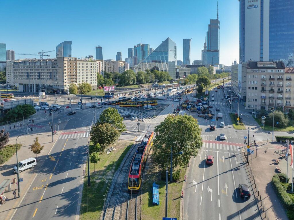 Widok na skrzyżowanie z tramwajem, samochodami i pieszymi w tle nowoczesne wieżowce i zieleń.
