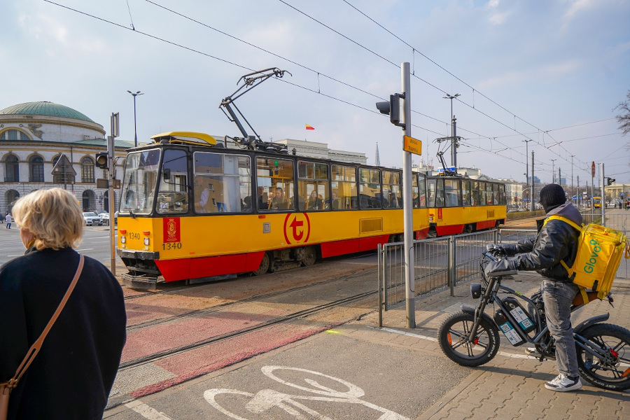 Tramwaj w żółto-czerwonych barwach zatrzymuje się na przystanku, obok stoi mężczyzna na rowerze z torbą dostawczą.