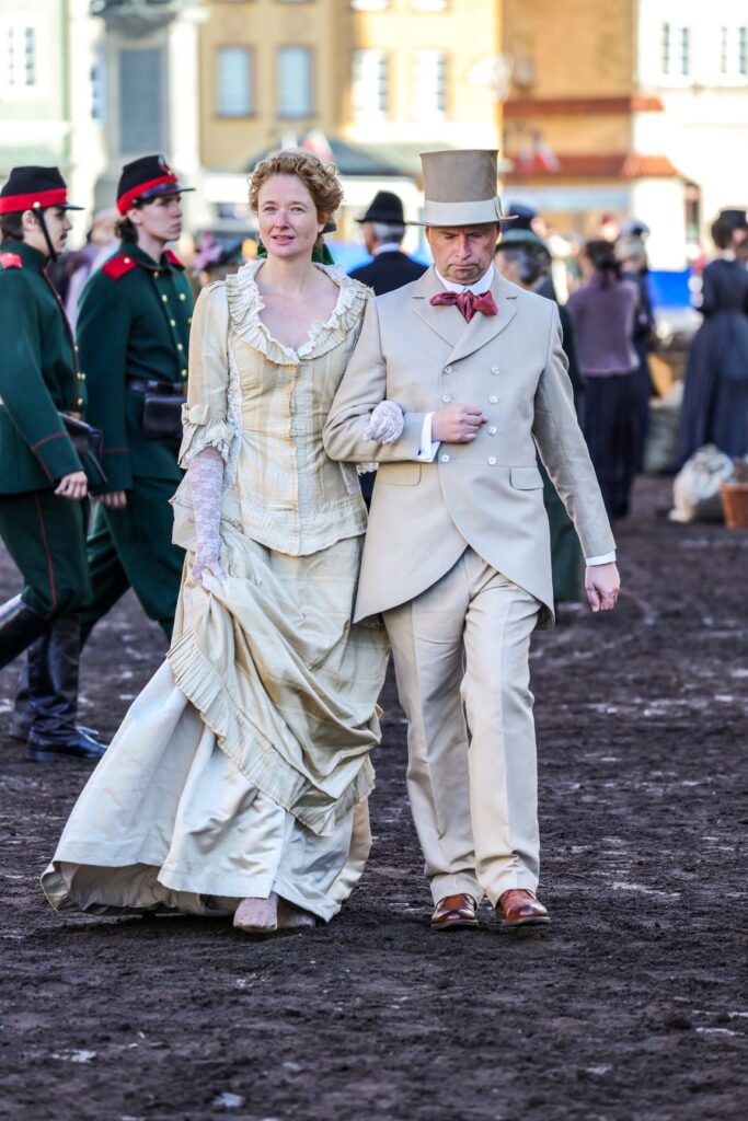 Mężczyzna w eleganckim garniturze prowadzi kobietę w stylowej sukni po błotnistej nawierzchni, w tle widoczni inni uczestnicy wydarzenia.