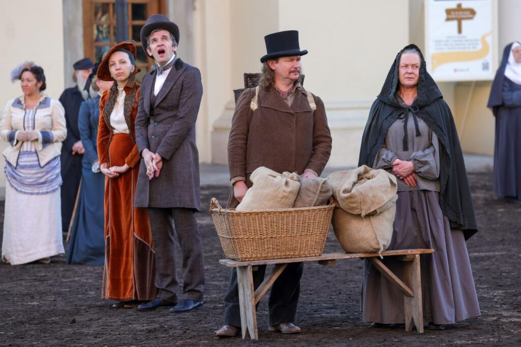 Grupa osób w strojach historycznych stoi na placu, mężczyzna w kapeluszu wyraża emocje, a obok stoi kosz z workami.