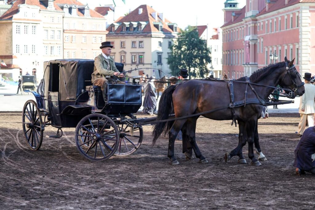 Mężczyzna w kapeluszu prowadzi powóz zaprzęgnięty w konia na tle zabytkowych budynków.
