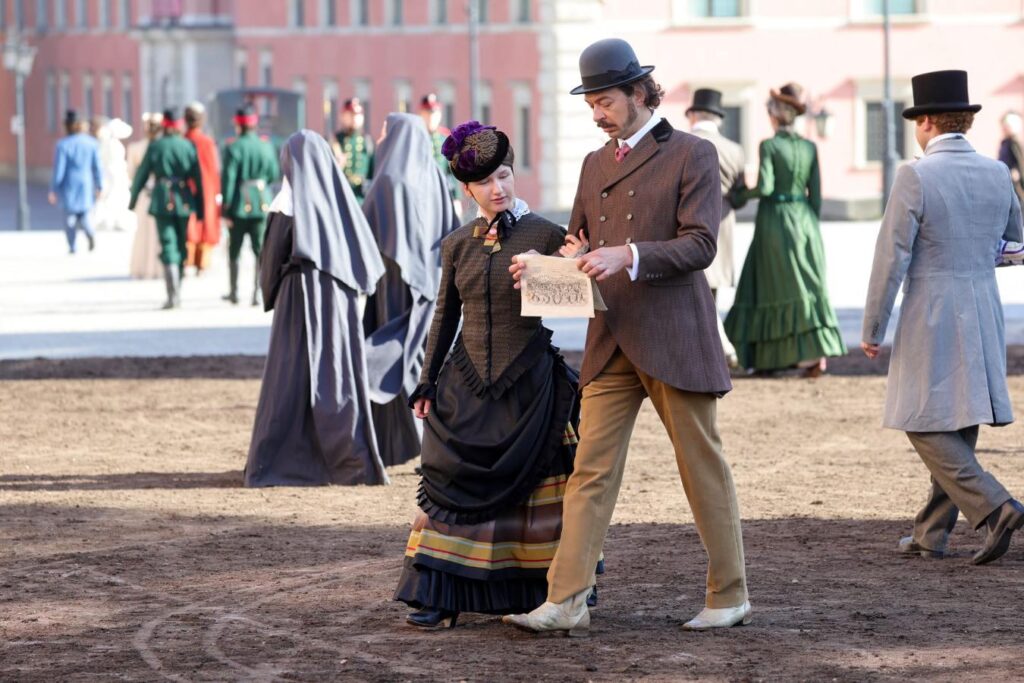 Mężczyzna w meloniku i eleganckim ubraniu rozmawia z kobietą w wiktoriańskiej sukni, trzymając w ręku kartkę. W tle widać postacie w historycznych strojach.