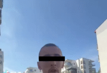 Mężczyzna w czarnej koszulce biega po osiedlu mieszkalnym, trzymając telefon w ręku i zwracając się do kamery. W tle nowoczesne budynki.