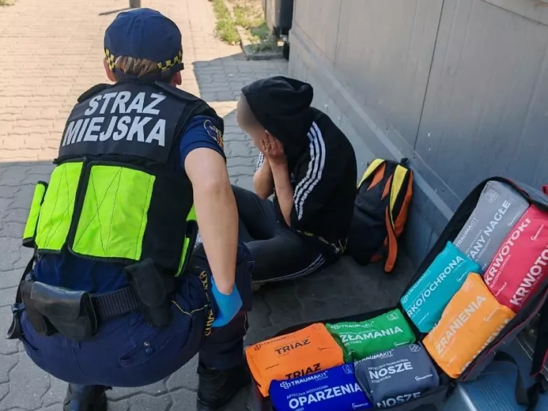 Stróż prawa w kamizelce z napisem "STRAŻ MIEJSKA" rozmawia z młodym człowiekiem siedzącym na chodniku, obok znajdują się torby z materiałami pierwszej pomocy.
