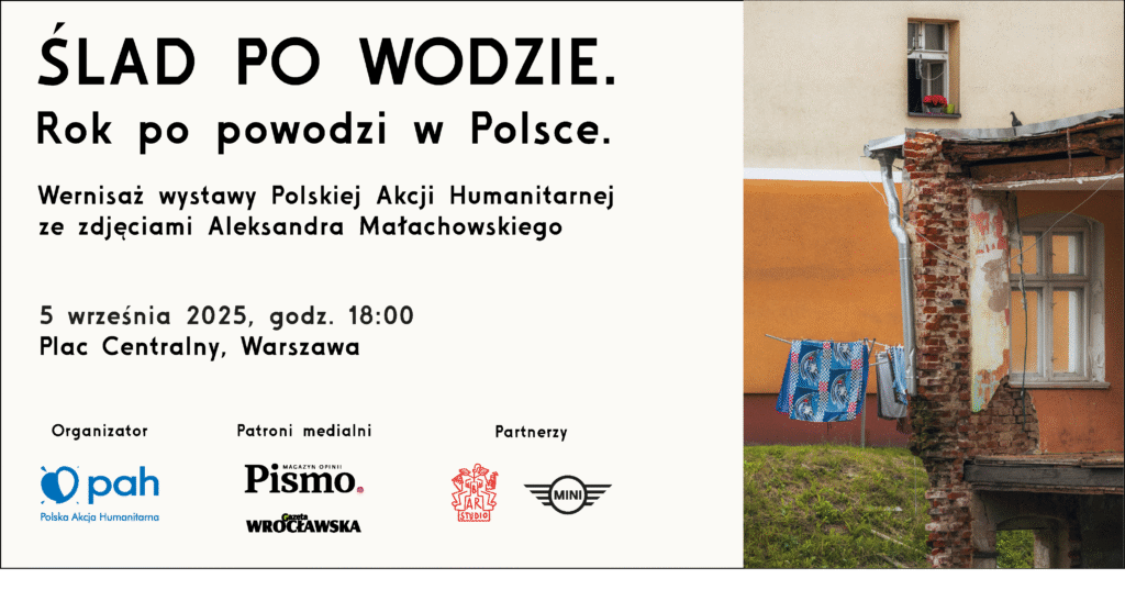 Wernisaż wystawy "Ślad po wodzie" z plakatem informacyjnym na tle budynku z praniem na sznurze.