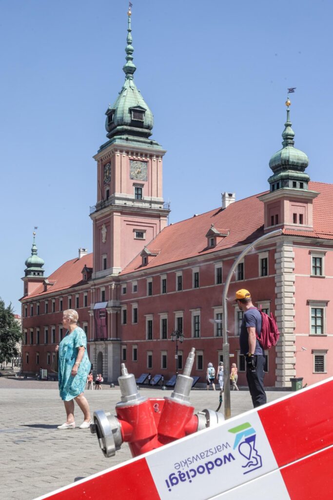 Kobieta w niebieskiej sukience spacerująca przed historycznym budynkiem z zielonymi kopułami, mężczyzna z plecakiem w tle.
