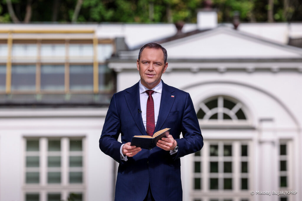 Mężczyzna w garniturze trzyma książkę i przemawia na tle nowoczesnego budynku z dużymi oknami.