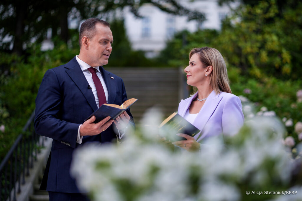 Mężczyzna w garniturze i kobieta w jasnej sukience trzymają książki, rozmawiając w ogrodzie pełnym kwiatów.