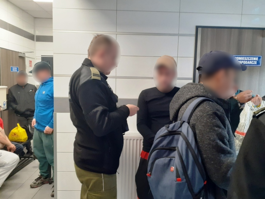Grupa osób w różnych ubraniach czeka w pomieszczeniu, podczas gdy funkcjonariusz w mundurze rozmawia z jednym z mężczyzn.