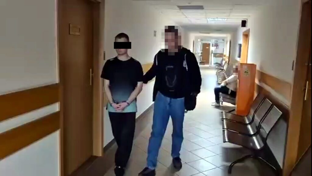 Mężczyzna w czarnej bluzie prowadzi osobę z rękami związanymi z tyłu przez korytarz w budynku.
