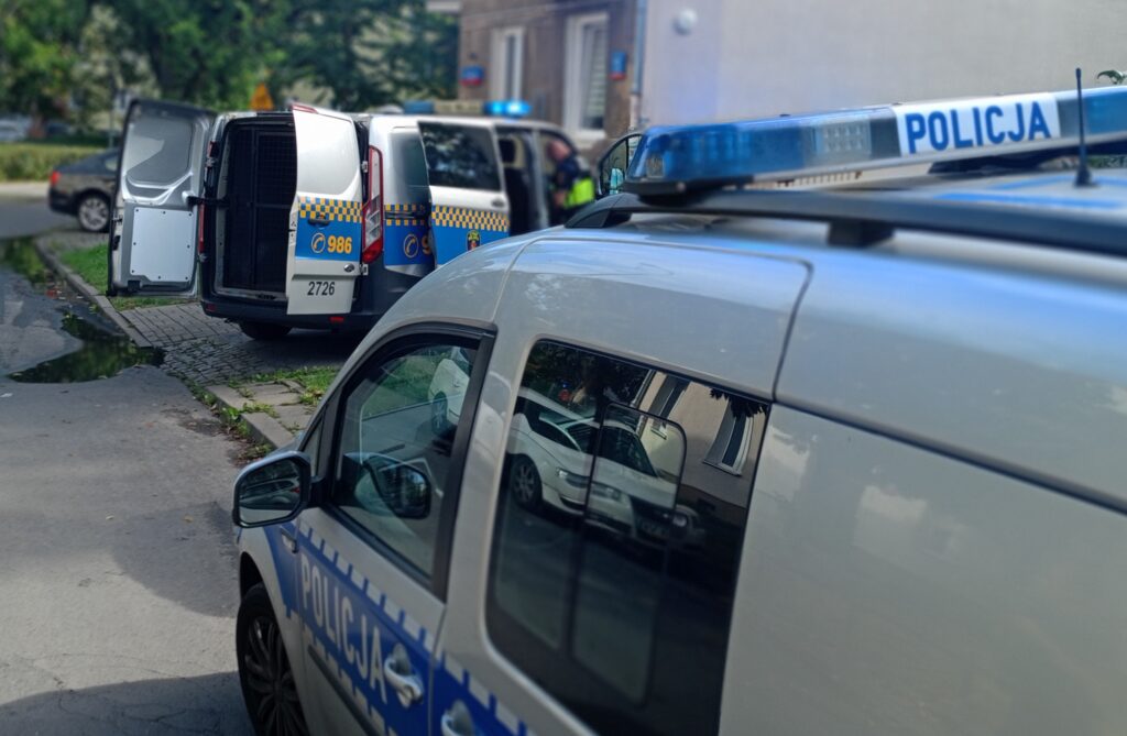 Dwa radiowozy policyjne zaparkowane na ulicy, jeden z otwartymi drzwiami, w tle widać budynki mieszkalne.