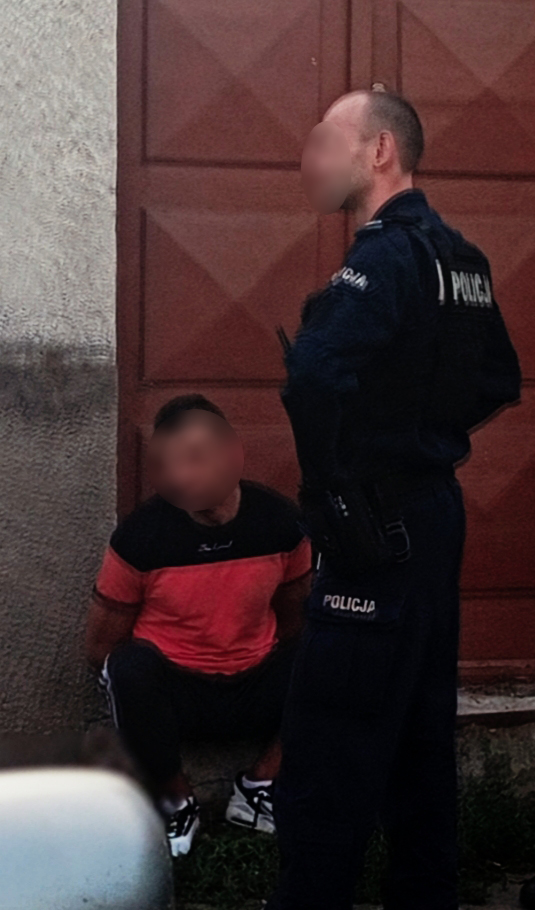 Policjant w mundurze stoi obok siedzącego mężczyzny w czerwonej koszulce przy drzwiach.