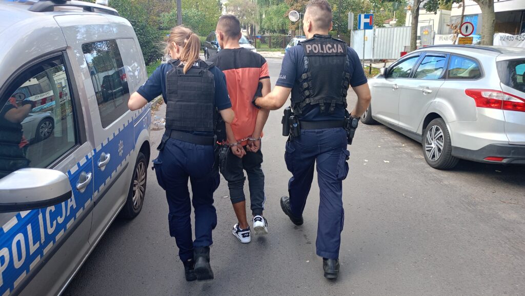 Dwóch policjantów prowadzi zatrzymanego mężczyznę w kajdankach na ulicy, obok radiowozu.