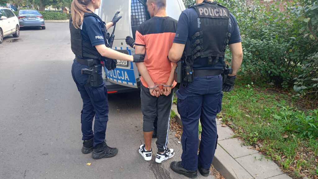 Mężczyzna w kajdankach zatrzymywany przez dwóch policjantów w pobliżu radiowozu na ulicy.