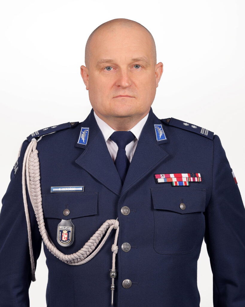 Mężczyzna w mundurze policyjnym z odznaczeniami, stojący na tle jasnej ściany.
