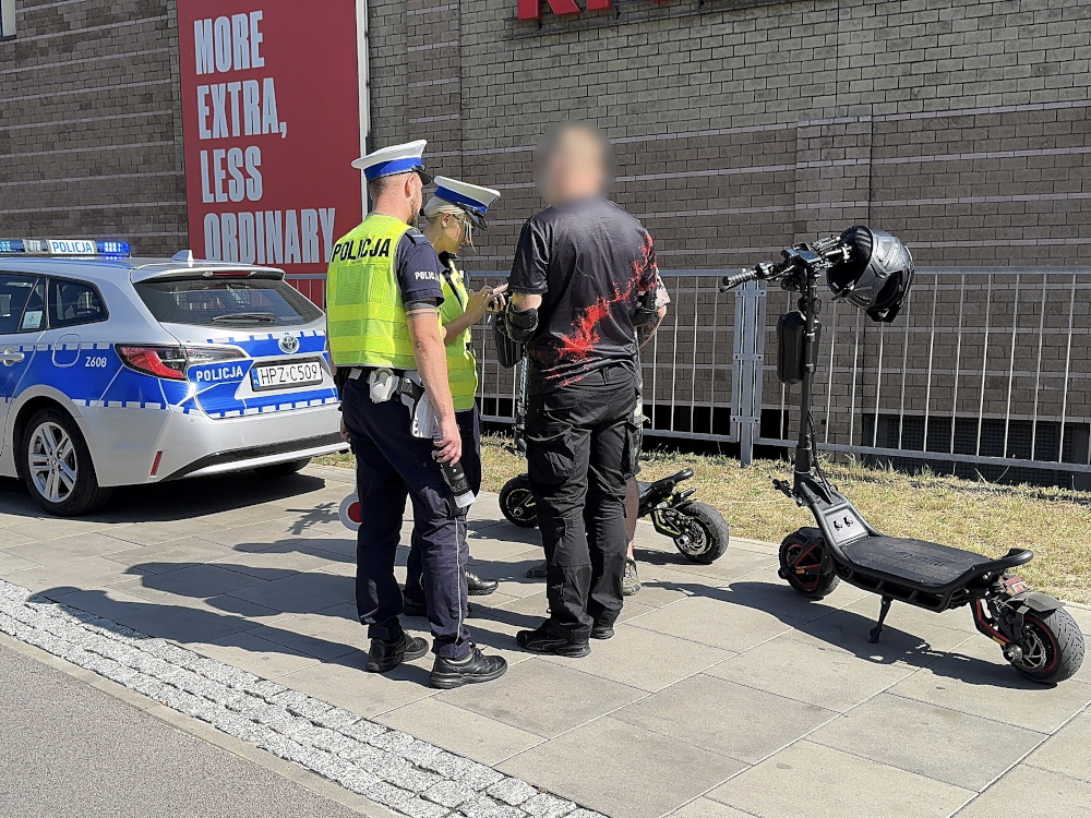 Policjanci w mundurach rozmawiają z mężczyzną przy dwóch hulajnogach elektrycznych obok radiowozu.