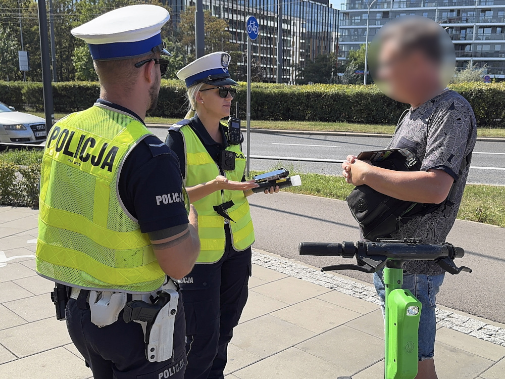 Policjantka w kamizelce odblaskowej rozmawia z mężczyzną stojącym obok zielonego hulajnogi na chodniku.