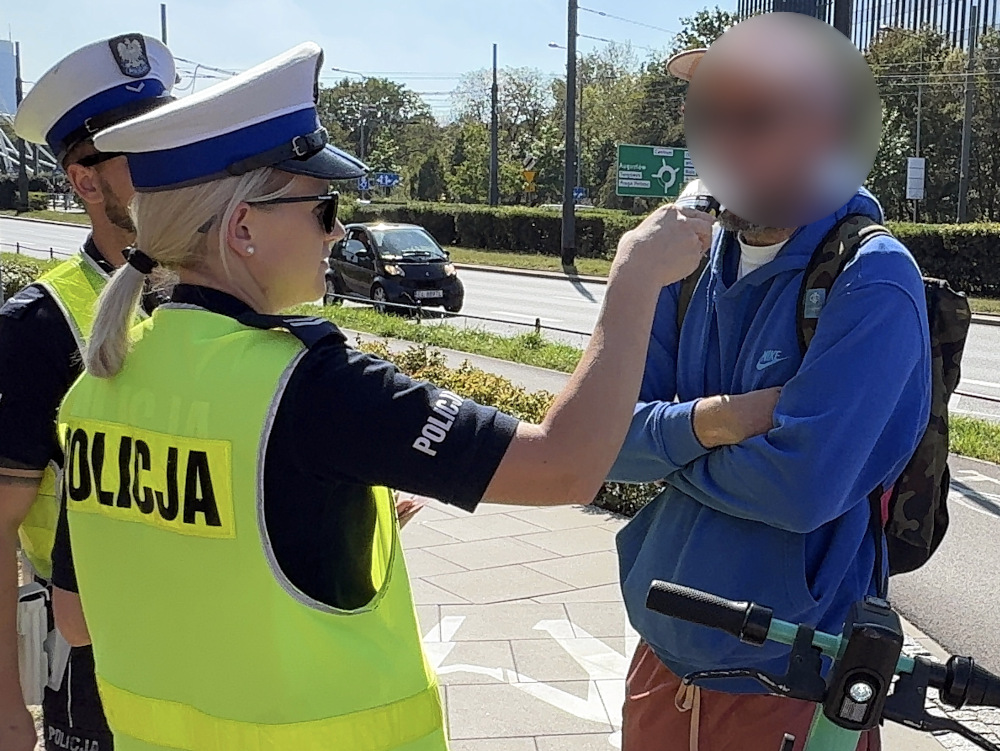 Policjantka w odblaskowej kamizelce kontroluje mężczyznę na ulicy, obok parkujących samochodów.