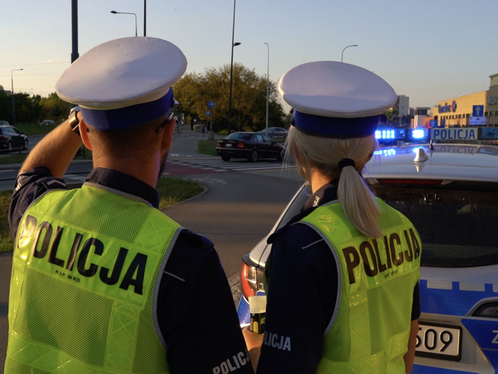 Dwójka policjantów w kamizelkach odblaskowych obserwuje ruch drogowy przy radiowozie z włączonymi sygnałami świetlnymi.