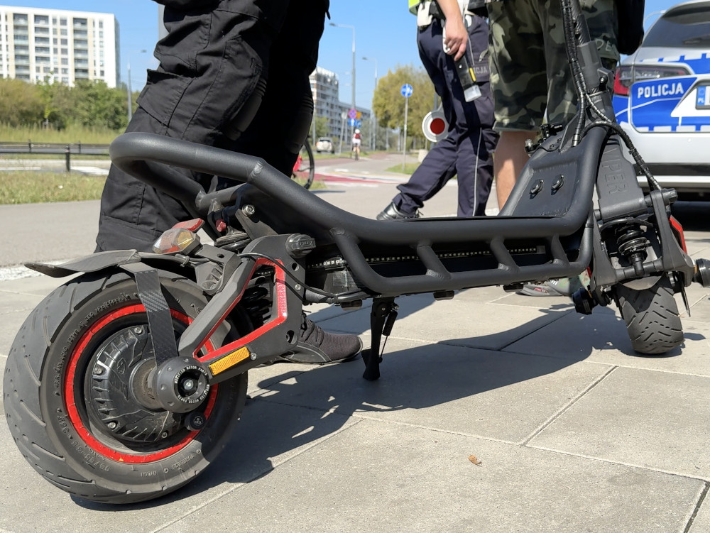 Policyjny funkcjonariusz sprawdzający elektryczną hulajnogę zaparkowaną na chodniku w miejskim otoczeniu.
