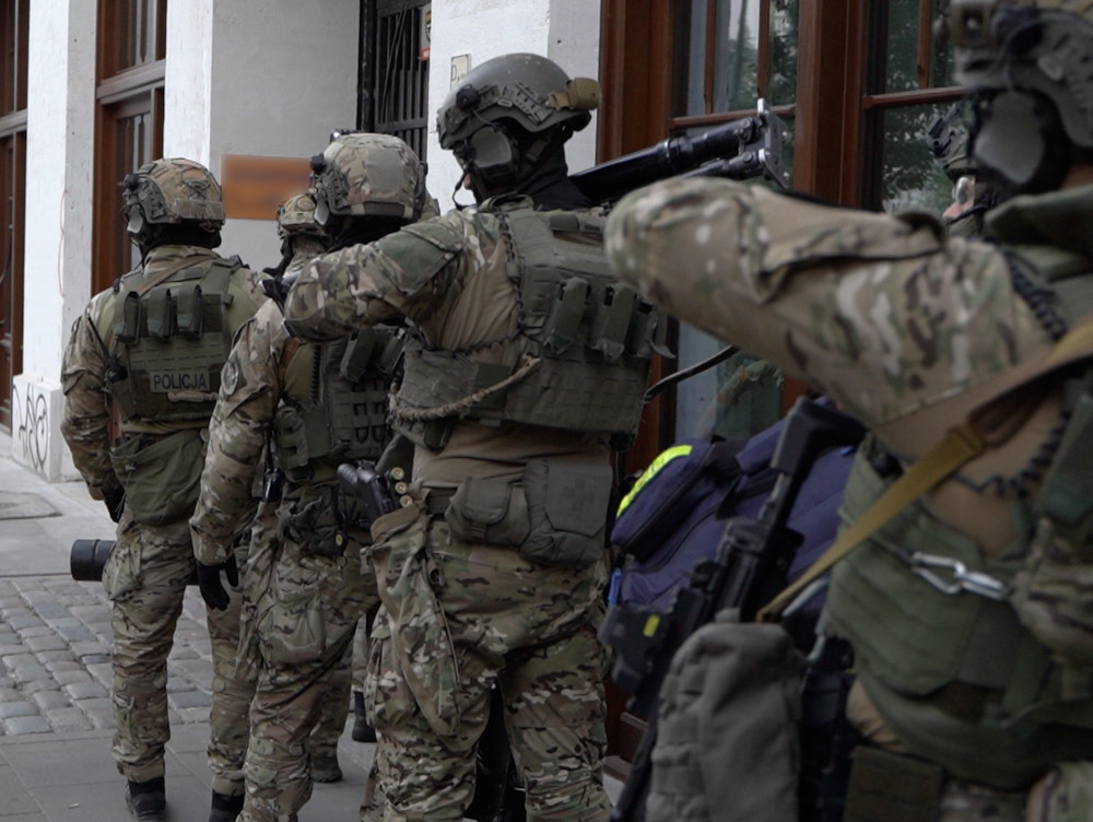 Grupa uzbrojonych funkcjonariuszy policji w mundurach taktycznych stoi w kolejce przed budynkiem z dużymi oknami.